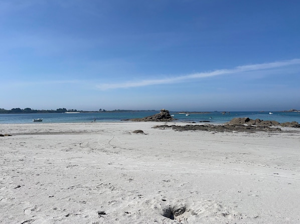 Une plage de sable parsemée de rochers, avec une eau calme et des bateaux visibles au loin.