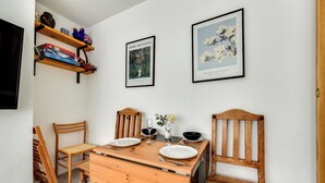 Ein Essbereich mit einem Holztisch, gedeckt für zwei Personen, zwei Stühlen, einer kleinen Kommode und zwei gerahmten Pflanzenbildern an der Wand.
