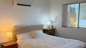 Une chambre à coucher comprenant un lit, une lampe de chevet, une petite table avec une lampe, une fenêtre et un appareil de climatisation.