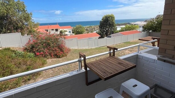 Un balcon avec une table en bois et deux tabourets blancs, donnant sur une zone côtière avec des maisons et une clôture.
