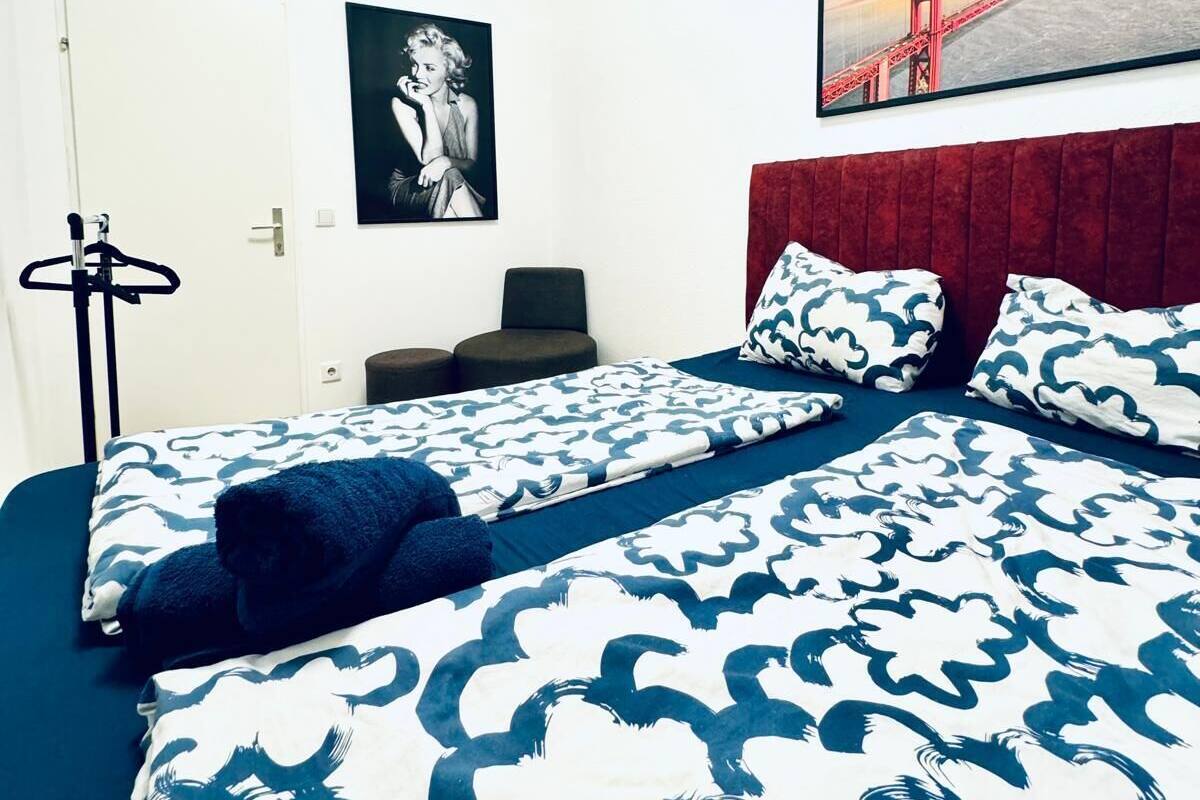 Ett hotellrum med en säng som har ett blått och vitt vågmönster, en röd sänggavel, en inramad bild av en bro och ett svartvitt porträtt på väggen.