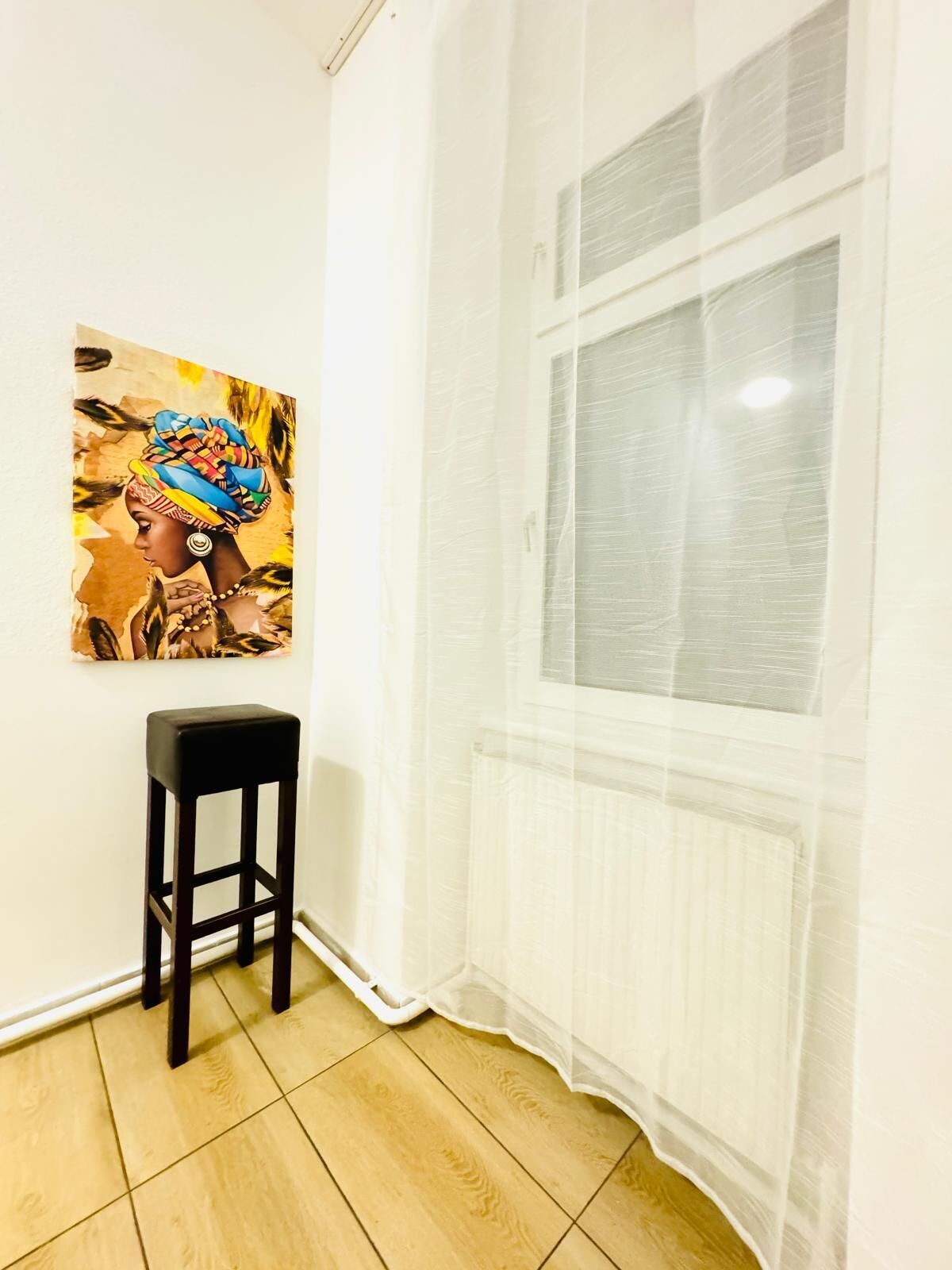 Ein Zimmer mit Fenster, Heizkörper, Holzboden und einem schwarzen Hocker.