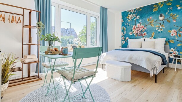 Un dormitorio con una ventana grande, una cama, una silla, una mesa y una pared decorada con un mural floral.