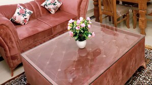 A living room with a red sofa, a glass coffee table with a flower vase, a dining table with wooden chairs, and an air conditioning unit on the wall. - Fortune Home Service Apartment E-28 2B (New Delhi)