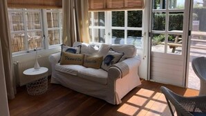 Ein Zimmer mit einer Couch, einem runden Tisch und einem Stuhl. Es gibt Fenster mit Jalousien und eine Tür, die nach draußen führt.