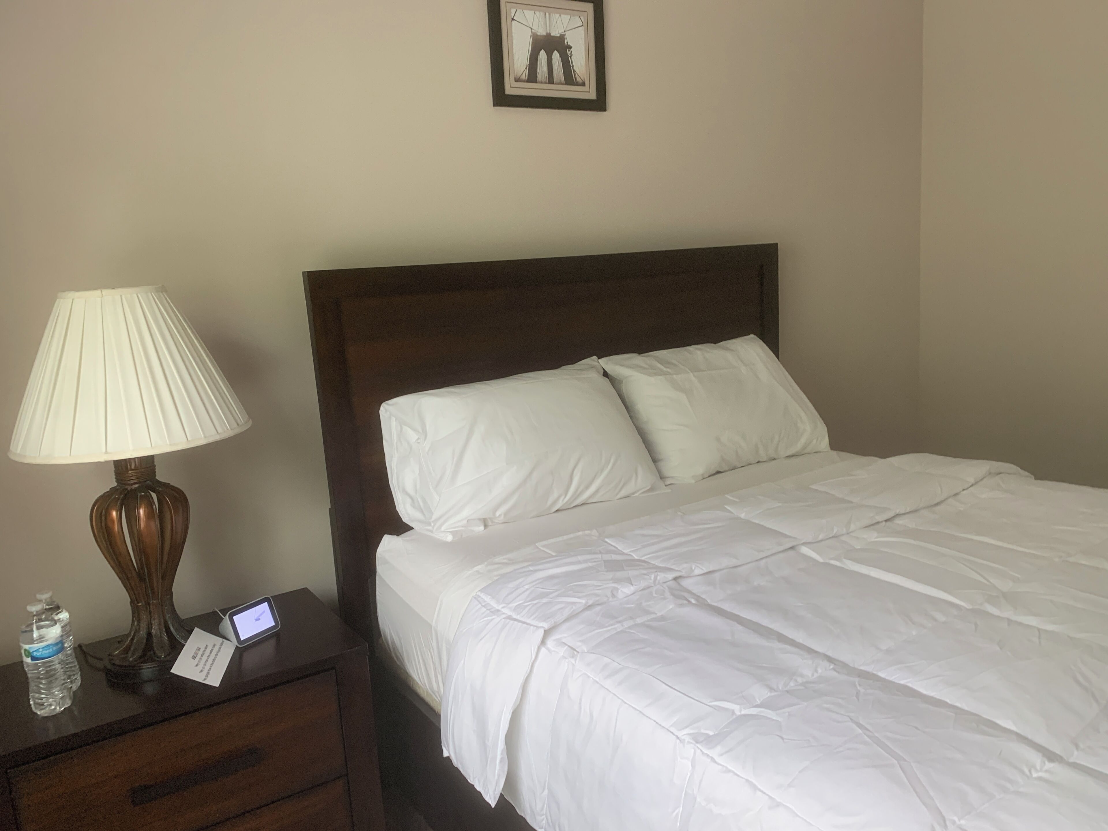 A hotel room with a bed, a nightstand with a lamp, a water bottle, and a digital clock.