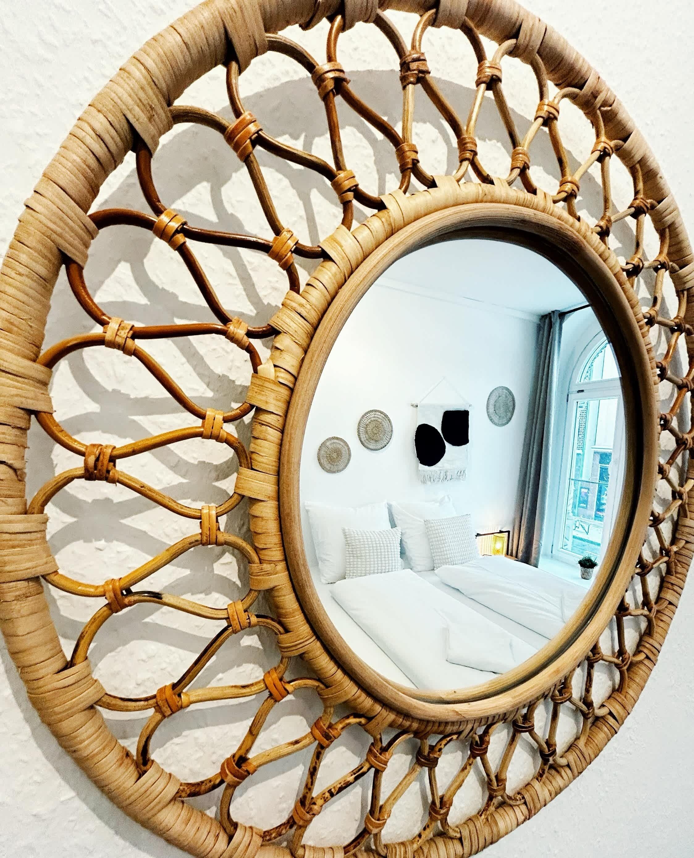 Een ronde spiegel met een geweven lijst, die een kamer weerspiegelt met een bed, kussens en een raam.
