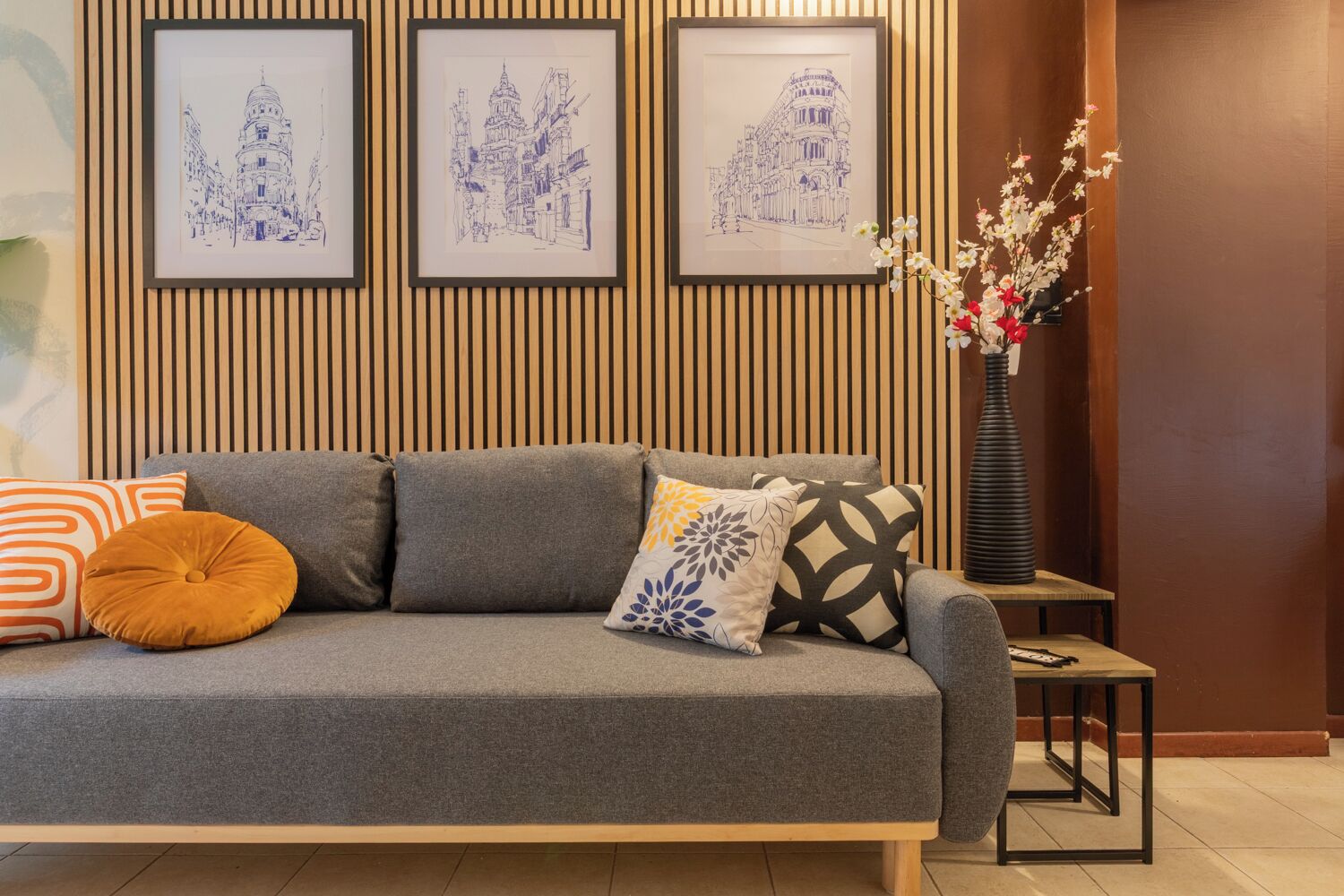 Ein graues Sofa mit gemusterten Kissen, ein kleiner Beistelltisch mit Blumen in einer Vase und drei blaue gerahmte Skizzen an der Wand.