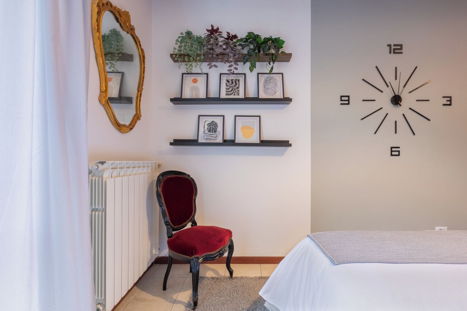 Una habitación con un radiador, una silla con cojín rojo, una cama con cubrecama blanco, un reloj de pared y estantes con obras de arte enmarcadas y plantas.