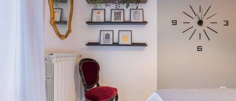 Una habitación con un radiador, una silla con cojín rojo, una cama con cubrecama blanco, un reloj de pared y estantes con obras de arte enmarcadas y plantas.