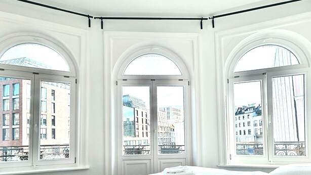 A hotel room with a large bed, three arched windows, and a view of city buildings. - Central Apartment for up to 10 Guests (Hamburg)