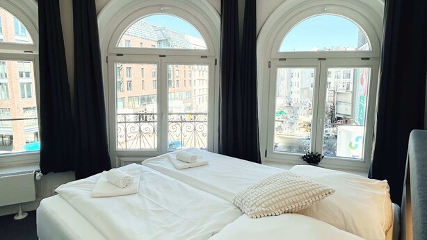Camera d'albergo con un letto grande, due finestre con tende nere, vista sui palazzi e una lampada a soffitto.