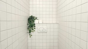 Un bagno piccolo rivestito di piastrelle bianche, con il water, un cestino, un porta carta igienica a muro e una pianta in vaso.