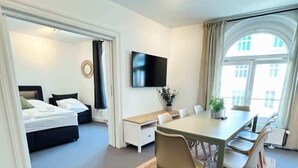Una moderna camera d'albergo con un letto, un'area pranzo con tavolo e sedie, una televisione a schermo piatto montata a parete e un'ampia finestra che offre luce naturale.