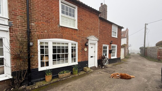A brick house with a white door, a window with a white frame, and a dog lying on the ground. - York Cottage (Reydon)