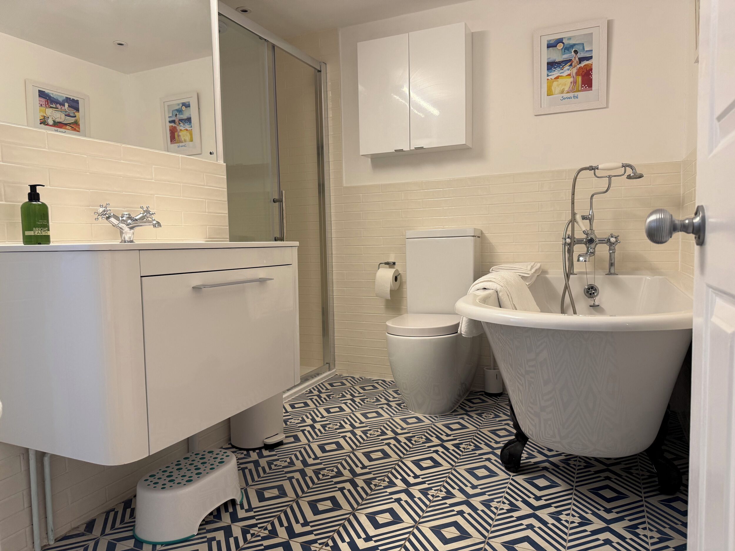 A bathroom with a white sink, toilet, and bathtub, patterned floor tiles, and a small step stool.