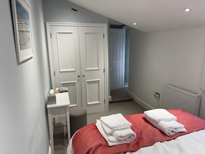 A bedroom with a bed, two white towels, a desk, a chair, a radiator, and a door leading to a staircase. - York Cottage (Reydon)