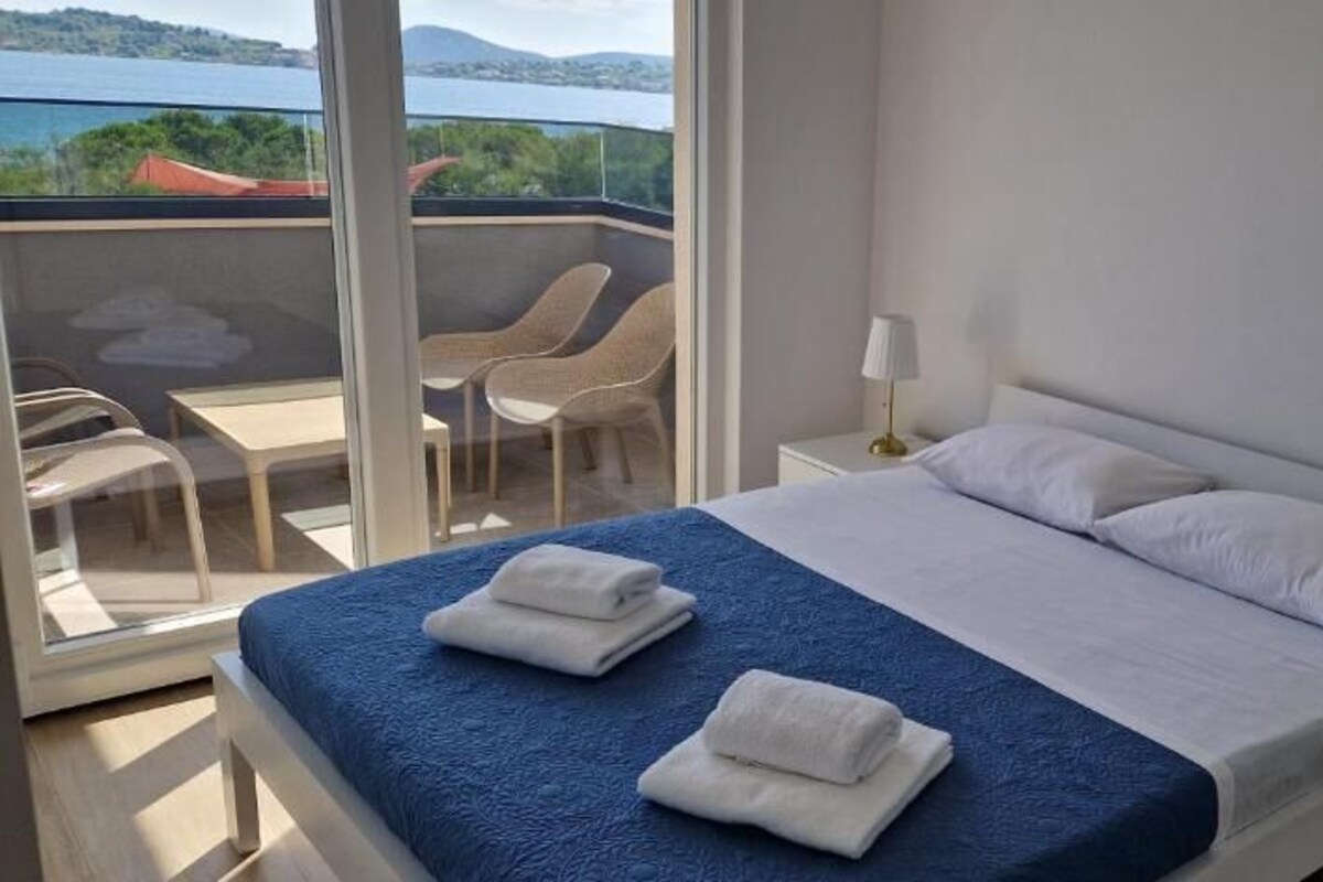 A bedroom with a bed, two bedside tables, a window with a view, and a balcony with chairs.