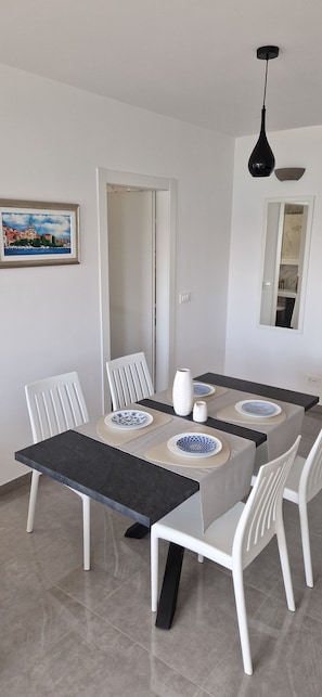 A dining area with a table set for four, white chairs, a black pendant light, a framed cityscape painting, and a mirror reflecting a bathroom. - Mare Anima Apartments (Srima)