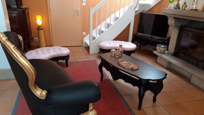 Un salon avec un fauteuil noir et doré, un banc rose rembourré, une table basse en bois, une cheminée et un escalier.