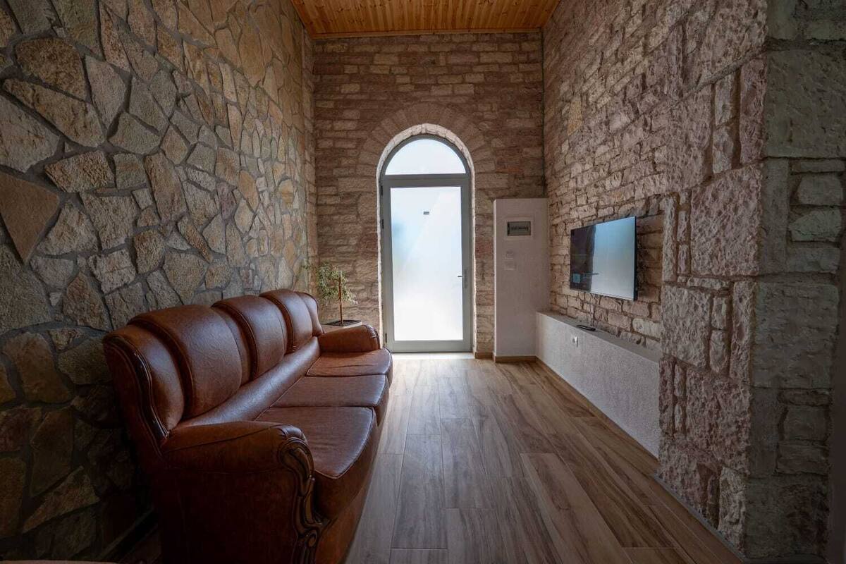 Una habitación con paredes de piedra y techo de madera, un sofá de cuero, un televisor montado en la pared y una puerta que da al exterior.