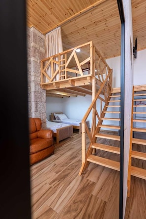 Una habitación con cama rústica en entrepiso, sofá, escalera de madera y pared de piedra.