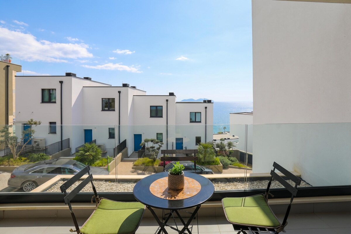 A balcony with a table and chairs, overlooking a residential area with buildings and a view of the sea.