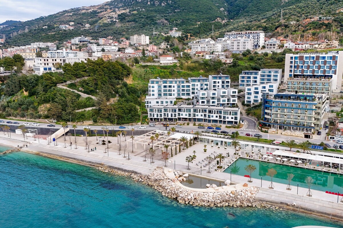 A coastal hotel with multiple buildings, a swimming pool, and a promenade along the waterfront.