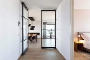 A modern hotel room with a wooden floor, a bed with white linens, a bedside table with a lamp, and a view through a glass door to a dining area with a table and chairs. - Luxury North Sea Retreat With Garage (Egmond aan Zee)