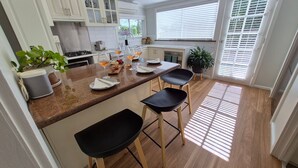 A modern kitchen with a central island, bar stools, and a view of the outdoors through large windows. - Finchy Cottage - Your charming Beechworth cottage where heritage character meets modern com (Beechworth)