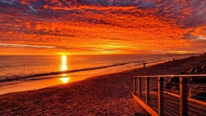 A beach with a sunset, a pier in the distance, and a fence along the shore.