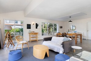 A spacious living area with a sofa, chairs, a dining table, and a large window providing a view of the outdoors. - Maison des Orchidées just 100m from the beach (La Tranche-sur-Mer)