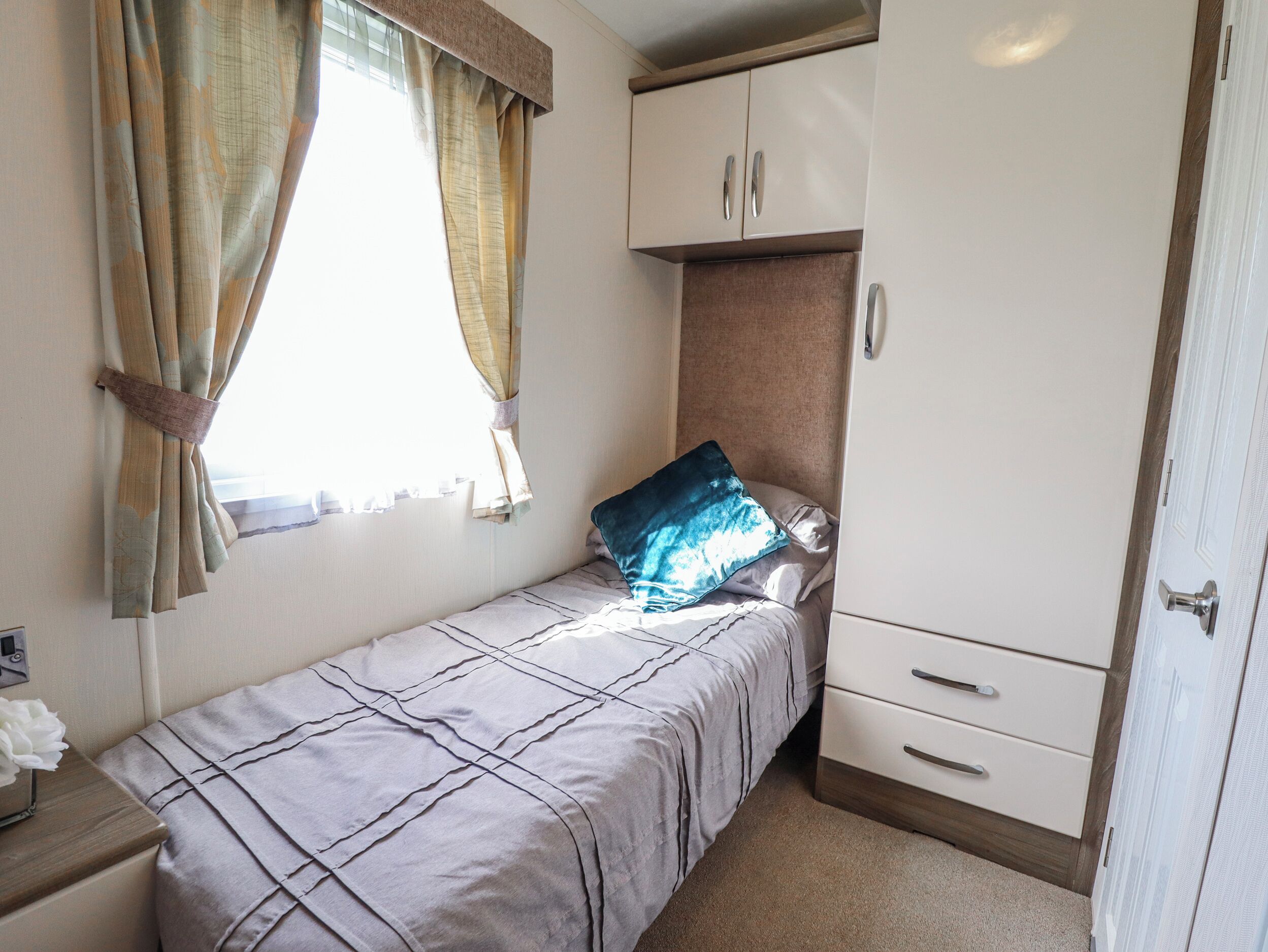 Un lit simple avec une couvre-lit à motifs, une table de chevet avec une lampe, une armoire à deux portes et une fenêtre avec des rideaux.