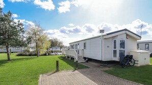 A mobile home with a paved driveway, a small attached garage, and a grassy yard with trees. - 13 The Lakes (Mablethorpe)