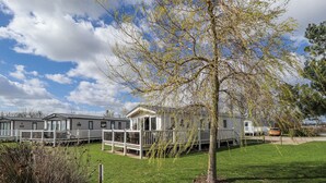 A row of mobile homes with a grassy lawn and a tree in the foreground. - 13 The Lakes (Mablethorpe)