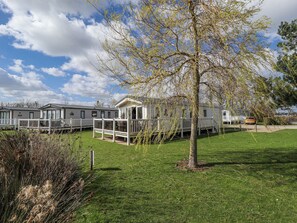 A row of mobile homes with a grassy lawn and a tree in the foreground. - 13 The Lakes (Mablethorpe)