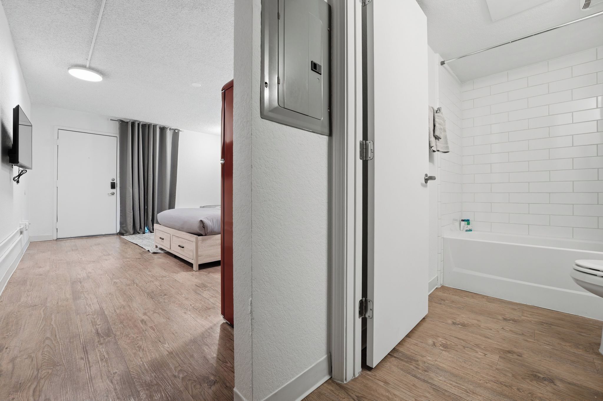 A hotel room with a wooden floor, a bed, a wardrobe, a bathroom with a bathtub, and a television mounted on the wall.