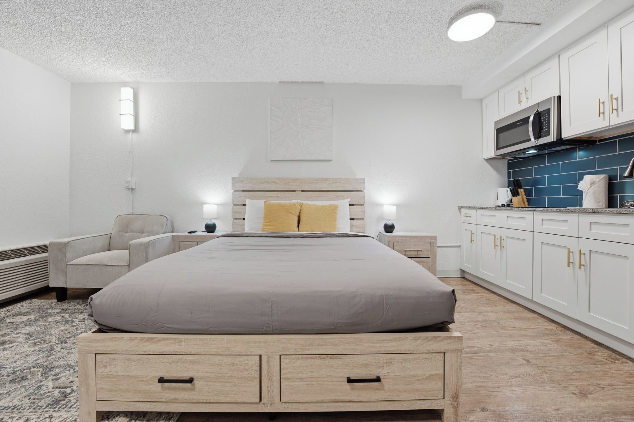 A modern bedroom with a bed, bedside tables, a chair, a built-in wardrobe, and a kitchenette.