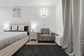 A bedroom with a bed, bedside table, chair, heater, and curtains. - Comfortable studio with updated amenities (Austin)