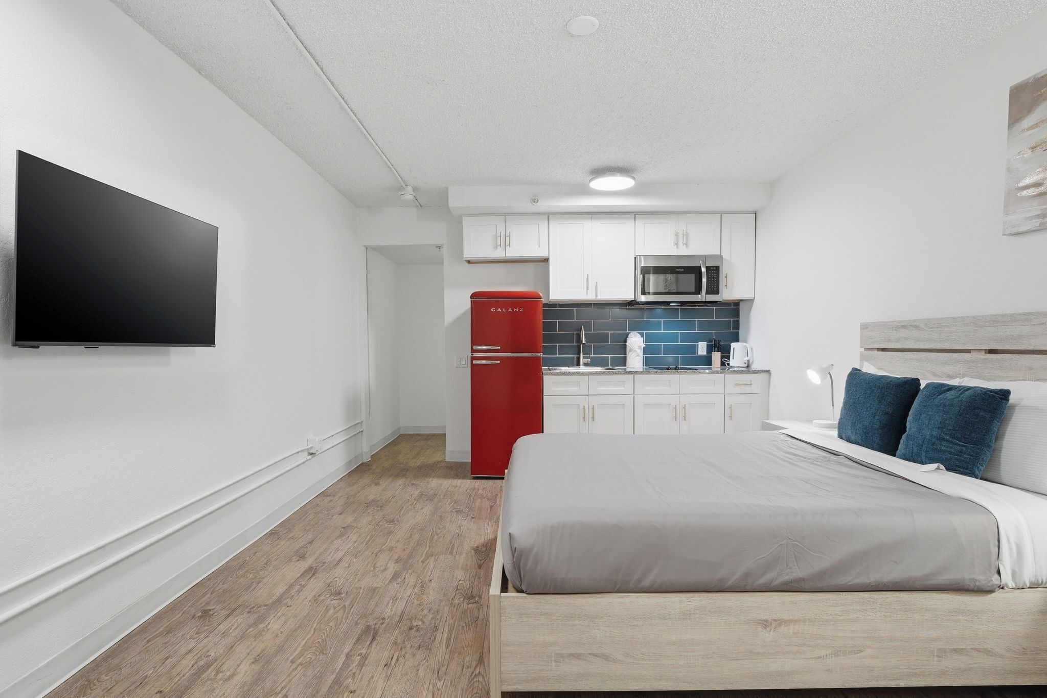 A bedroom with a bed, a TV, a red refrigerator, and a kitchen area with cabinets and a microwave.
