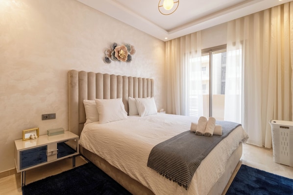 A modern bedroom with a large bed, a bedside table with a lamp, and a view of the city through the window.