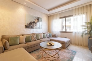 A living room with a sectional sofa, a glass coffee table, a large abstract painting, and a view of the outside through sheer curtains.