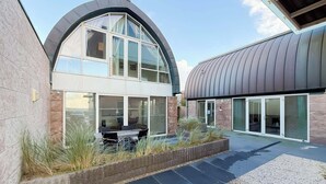 Un edificio moderno con techo curvo, amplias ventanas y un patio con mesas al aire libre.