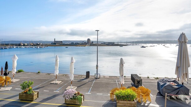 Blick auf das Wasser mit einem Hafenbecken, Sonnenschirmen, Sitzgelegenheiten und einer Straßenlaterne.