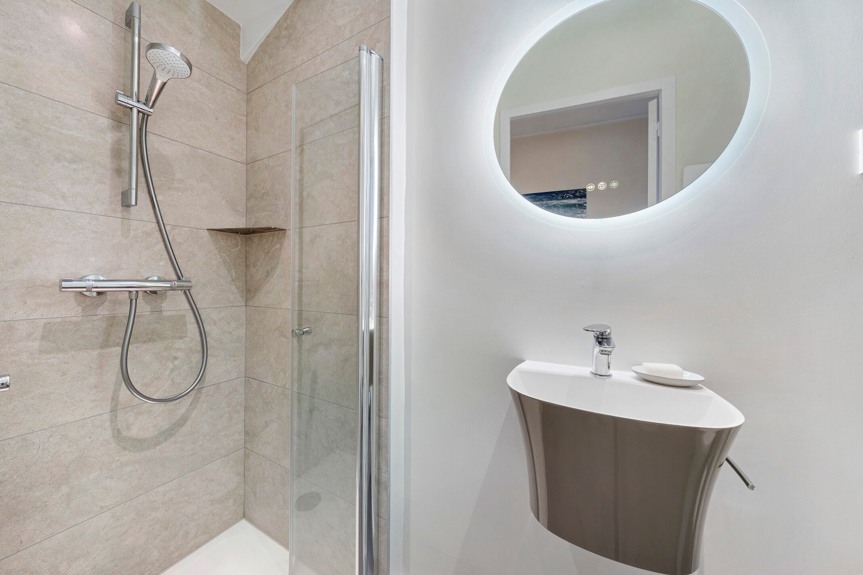 Un baño moderno con cabina de ducha de vidrio, un espejo redondo y un lavabo sobre pedestal.
