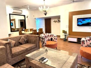 A living room with a sofa, two armchairs, a glass coffee table, a TV, and a dining area in the background. - Fortune Home Service Apartment E-28 1A (New Delhi)