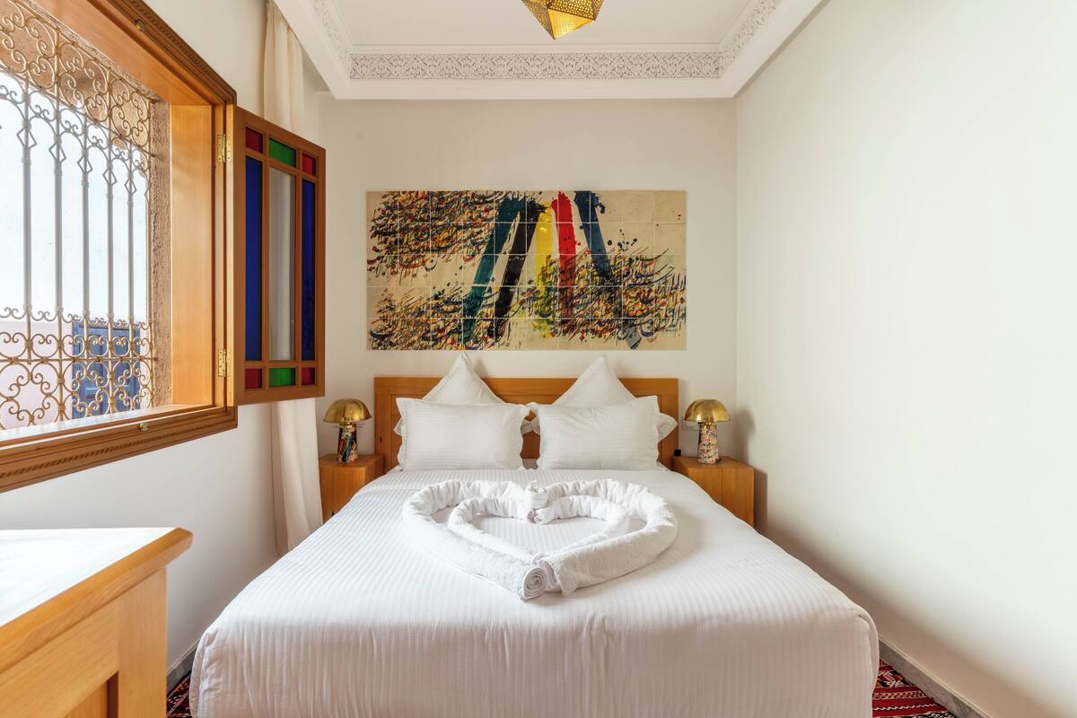 A hotel room with a bed, a window with decorative bars, a painting on the wall, and a heart-shaped towel on the bed.