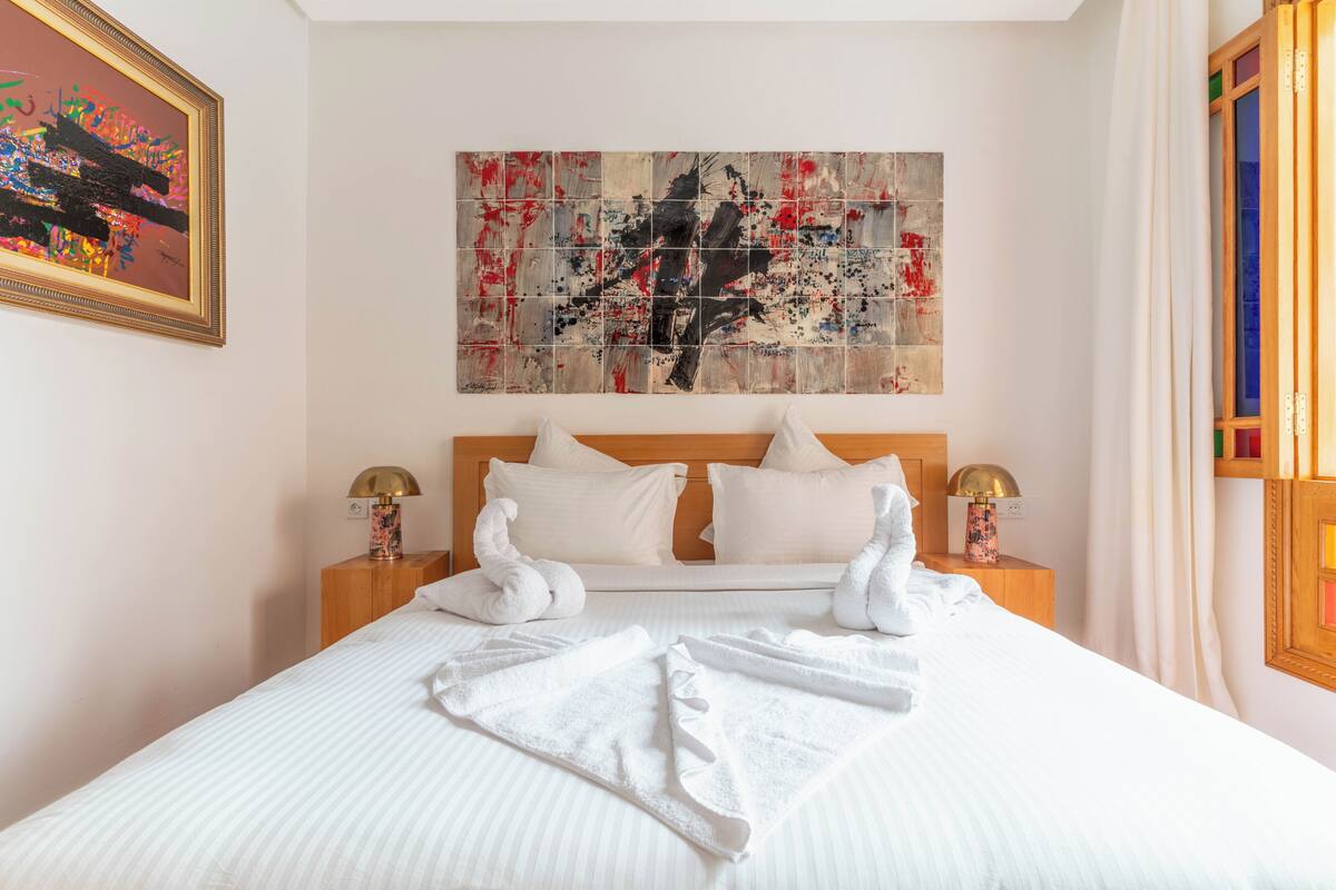 A hotel room with a large bed, two bedside tables, a headboard, a framed abstract painting, and a window with curtains.