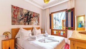 A hotel room with a large bed, a headboard, a nightstand, a lamp, a painting, a window with colorful shutters, and a wooden dresser. - Riad House of Calligraphy Marrakech (Marrakech)