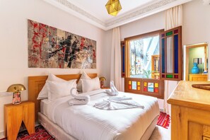 Habitación de hotel con una cama grande, cabecera, mesita de noche, lámpara, un cuadro, ventana con contraventanas coloridas y un aparador de madera.